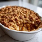 Close-up of a freshly baked Apple Crisp in a white dish, showing bubbling fruit filling and a crunchy oat topping.