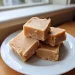 A stack of four creamy squares of homemade Peanut Butter Fudge resting on a white plate.