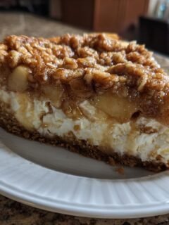 A delicious slice of Apple Crisp Cheesecake on a white plate, showcasing layers of creamy cheesecake, apple filling, and crisp topping.