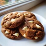 Four rich, brown Bakery Style Gingerbread White Chocolate Chip Cookies stacked on a white plate near a window.