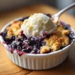 Close-up of a warm, bubbling Blueberry Cobbler topped with a scoop of vanilla ice cream and a spoon ready to dig in.