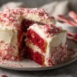 A slice of red velvet Candy Cane Cake with white frosting and crushed candy canes.