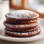 A close-up of a stack of three rich Chocolate Pancakes dusted generously with white powdered sugar.