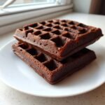 Two perfectly cooked, dark brown Chocolate Waffles stacked on a white plate near a window.