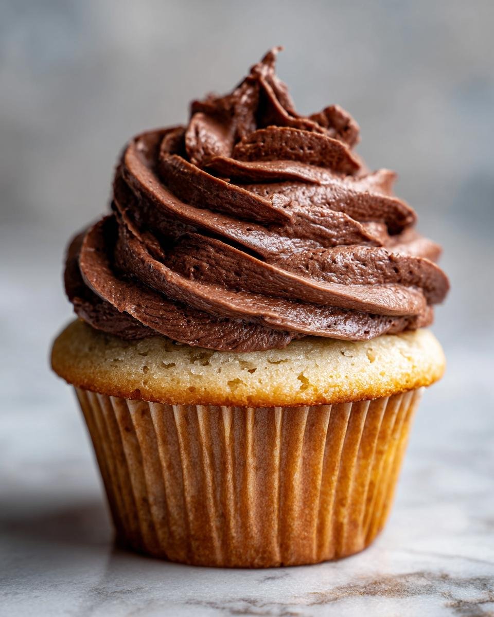 Close-up of a vanilla cupcake topped with a tall swirl of rich, dark brown Nutella Frosting.