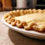 Close-up of a beautifully crimped, unbaked flaky pie crust edge sitting in a white pie dish.