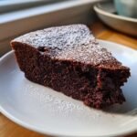 A decadent slice of Flourless Chocolate Cake dusted with white powdered sugar, served on a white plate.