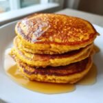 A tall stack of three golden-orange Fluffy Pumpkin Pancakes drizzled generously with maple syrup on a white plate.