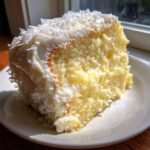 A close-up of a moist slice of Lemon Cake With Coconut Frosting, topped with shredded coconut.