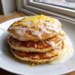 A stack of three fluffy Lemon Pancakes drizzled with white glaze and topped with bright yellow lemon zest.