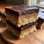 Two No Bake Chocolate Peanut Butter Bars stacked on a wooden board, showing the crust, peanut butter layer, and chocolate topping.