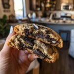 A hand holding two halves of a delicious Oreo Chocolate Chip Cookie, revealing chunks of Oreo cookies inside.