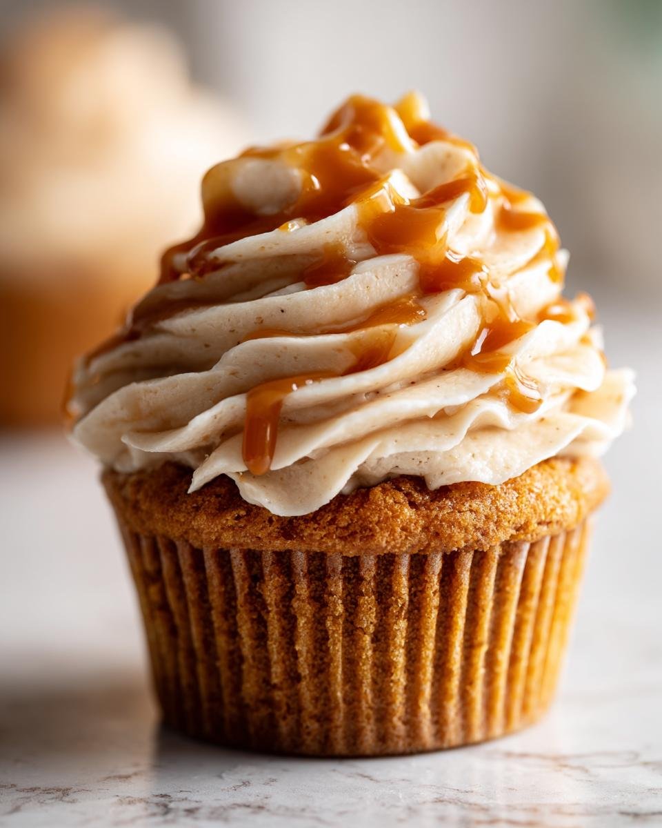 A close-up of a delicious Pumpkin Cupcake topped with swirled Caramel Cream Cheese Frosting and drizzled with caramel sauce.