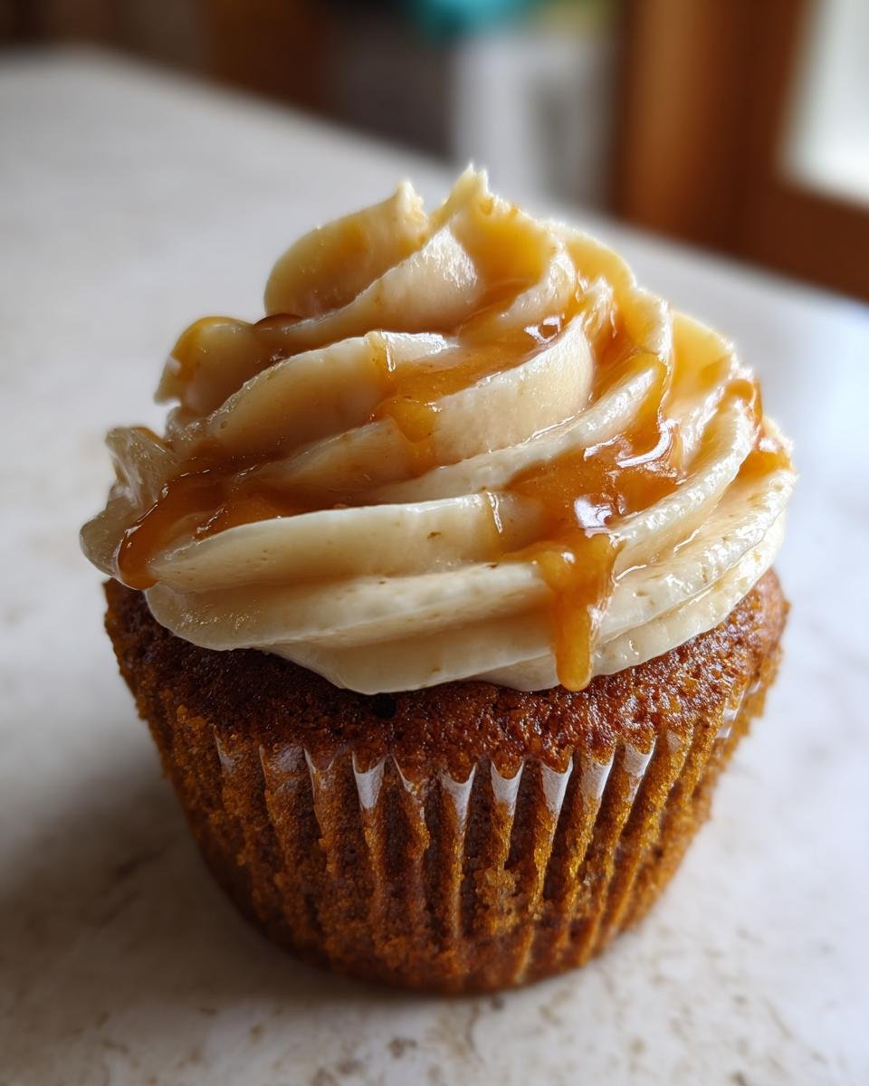 A close-up of a delicious pumpkin cupcake topped with caramel cream cheese frosting and drizzled with caramel sauce.