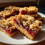 Four squares of homemade Raspberry Crumble Bars stacked on a white plate, showing a thick jam filling and crumbly topping.