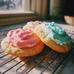 Close-up of soft batch frosted sugar cookies, one with pink frosting and another with blue frosting, on a cooling rack.