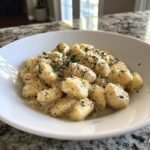 A close-up of The Best Cacio E Pepe Gnocchi coated in creamy cheese sauce, black pepper, and parsley in a white bowl.