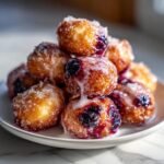 Close-up of a stack of golden-brown Blueberry Donut Holes coated in sugar and drizzled with white glaze.