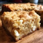 A close-up shot of a chewy square of White Chocolate Blondies studded with melted white chocolate chips on a wooden board.