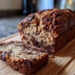 A close-up of freshly baked Chocolate Banana Bread loaf with one slice cut, showing melted chocolate chips throughout.