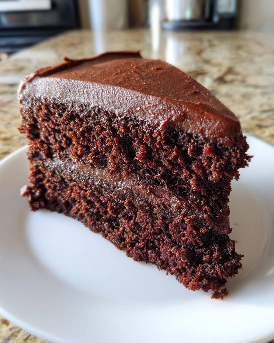 A close-up of a moist slice of Chocolate Cupcake Cake with thick chocolate frosting on a white plate.