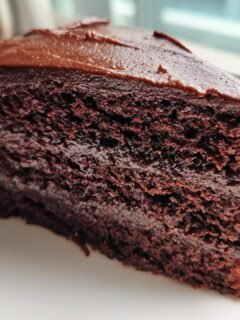 Close-up of a moist, dark slice of Chocolate Fudge Cake topped with thick chocolate frosting on a white plate.