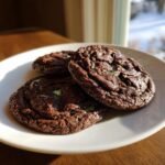 Three rich, dark Chocolate Mint Cookies with visible green mint chips stacked on a light plate.