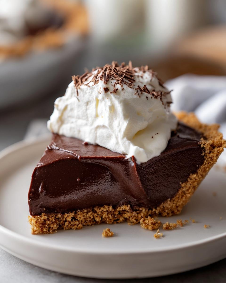 A decadent slice of Chocolate Pudding Pie with a thick, glossy filling and graham cracker crust, topped with whipped cream and chocolate shavings.