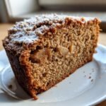 A close-up of a moist slice of Cinnamon Apple Cake, featuring visible apple chunks and a dusting of powdered sugar on top.