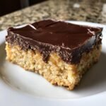 A close-up view of a square slice of Peanut Butter Bars with a thick, shiny chocolate topping on a white plate.