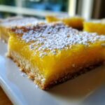 A close-up, tempting shot of a square Lemon Bar with a thick, bright yellow filling and shortbread crust, dusted with powdered sugar.