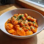 A white bowl filled with gnocchi-like dumplings coated in a rich, creamy tomato sauce, garnished with parsley.