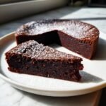 A slice of rich, dark Decadent Flourless Chocolate Cake dusted with powdered sugar, next to the remaining cake.