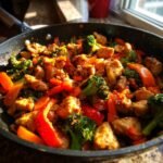 Close-up of Delicious Chicken Stir Fry With Veggies in a black skillet, featuring chicken pieces, broccoli, and red peppers.