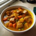 Close-up of a bowl filled with Delightful Tofu Curry, featuring cubes of tofu, carrots, potatoes, and peas in a rich, orange sauce.