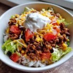 A close-up photo of Easy Beef Burrito Bowls featuring seasoned ground beef, rice, lettuce, tomatoes, cheese, and a dollop of sour cream.