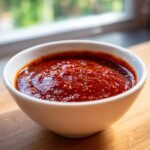 A close-up of rich, thick Easy Homemade Enchilada Sauce in a white bowl on a wooden surface.