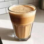 A glass filled with a thick, frothy Easy Peanut Butter Banana Oats Smoothie sitting on a white countertop.