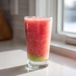 A tall glass filled with a vibrant Frozen Cucumber Watermelon Strawberry Mocktail, showing a layered effect with pink slush on top and green at the bottom.
