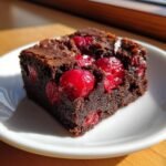 Close-up of a fudgy square of Cherry Brownies studded with bright red, whole cherries on a white plate.