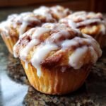 A close-up of a freshly baked Cinnamon Roll Muffin topped with thick white icing dripping down the sides.