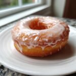 A single, perfectly glazed Vanilla Donuts resting on a white plate near a window.