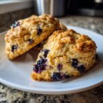 Close-up of two freshly baked, golden brown Blueberry Scones studded with juicy blueberries on a white plate.