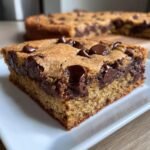 A thick, gooey square of Chocolate Chip Blondies served on a white plate with the rest of the pan blurred in the background.