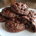A stack of rich, dark Hot Chocolate Cookies on a white plate, one broken open showing a gooey center.
