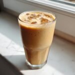 A tall glass filled with a creamy, frothy iced coffee Frappe and ice cubes, sitting on a white marble surface.