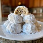 A stack of Irresistible Pecan Snowball Cookies heavily dusted in powdered sugar, with one cookie broken open showing the pecan filling.