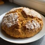 A golden brown, round loaf of Irresistible Vegan Soda Bread dusted with flour, resting on a white plate.