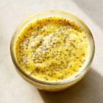 Overhead view of thick, bright yellow Lemon Poppy Seed Dressing Dairy Free speckled with black poppy seeds in a clear glass jar.