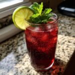 A tall glass filled with a bubbly, deep red Loaded Hibiscus Mocktail, garnished with a lime wheel and fresh mint sprigs.